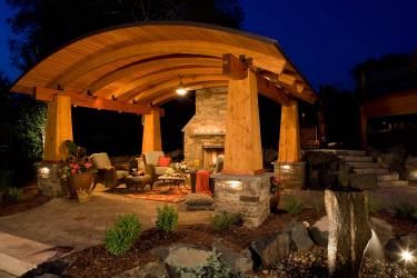 arched wooden outdoor structure sheltering patio furniture and fireplace at night