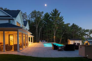 The moon shines bright in the sky over a pine forest and luxury lake house backyard with a swimming pool and porcelain tile patio.