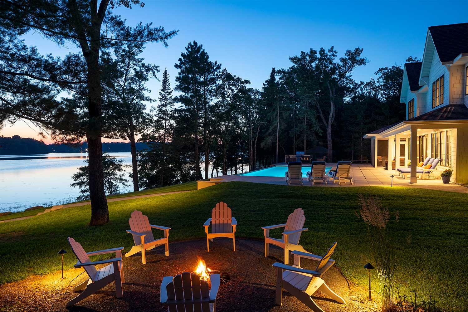 Northern Minnesota lakeshore landscape at dusk. A fire surrounded by a circle of white Adirondack chairs in the foreground, with the illuminated swimming pool and pool deck in the background.