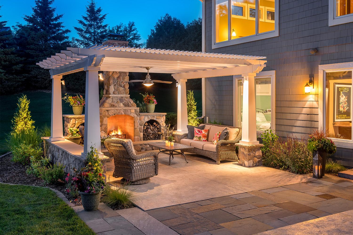 Outdoor living room patio with stone-faced fireplace underneath a white pergola at dusk