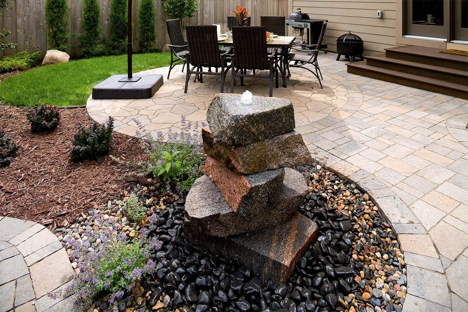 Stacked stone ornamental water fountain with dining patio and dog grass in the background.