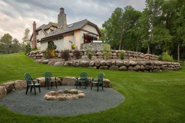 Glacial boulder retaining wall with shrubs and trees. In the foreground, a rustic campfire circle.